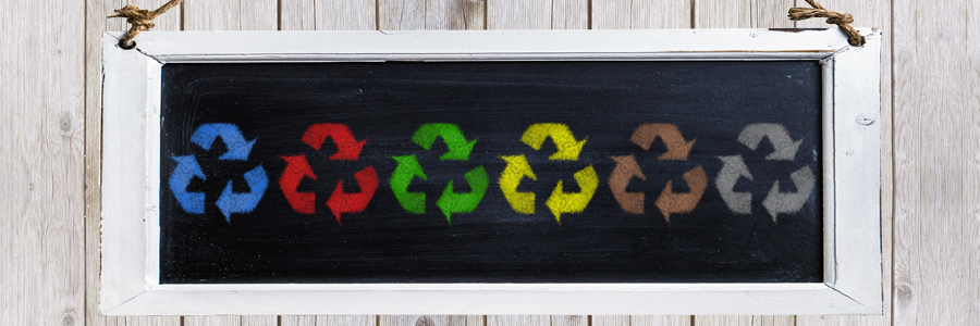 Imagem com fundo de madeira e um quadro negro no centro pendurado por uma corda, dentro do quadro seis símbolos da reciclagem rabiscados nas cores azul, vermelho, verde, amarelo, marrom e cinza.